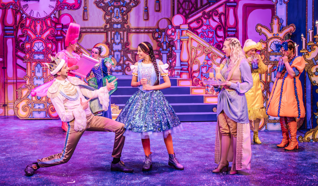 Photo of Cinderella wearing a sparkly sky-blue knee-length ball gown, in the centre of a gilded ballroom. She is smiling at the prince, wearing a golden crown and gold and lace formal wear, lunging and pointing at her.