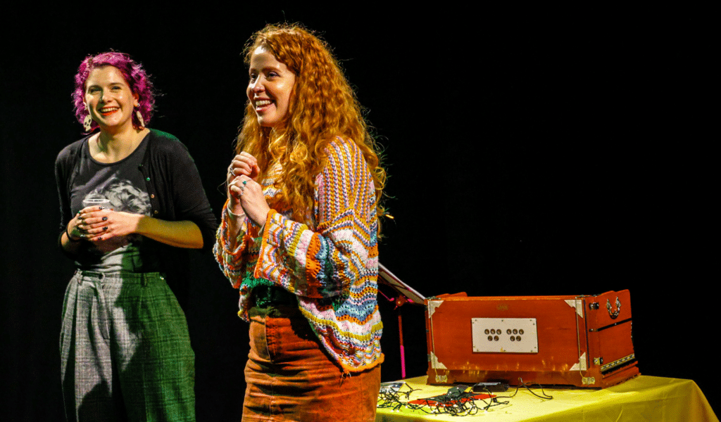 Photo of Katy Danbury and Naomi Turner stood on stage under theatre lighting, and smiling at an off-stage audience.