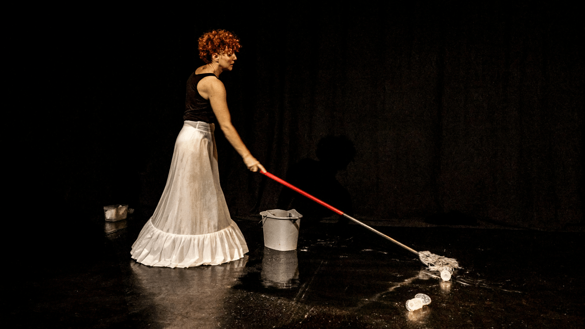 Photo of a performer in a black tank top and a white maxi skirt, alone in a black studio space. She is mopping the floor, where two breast pumps are spattered.