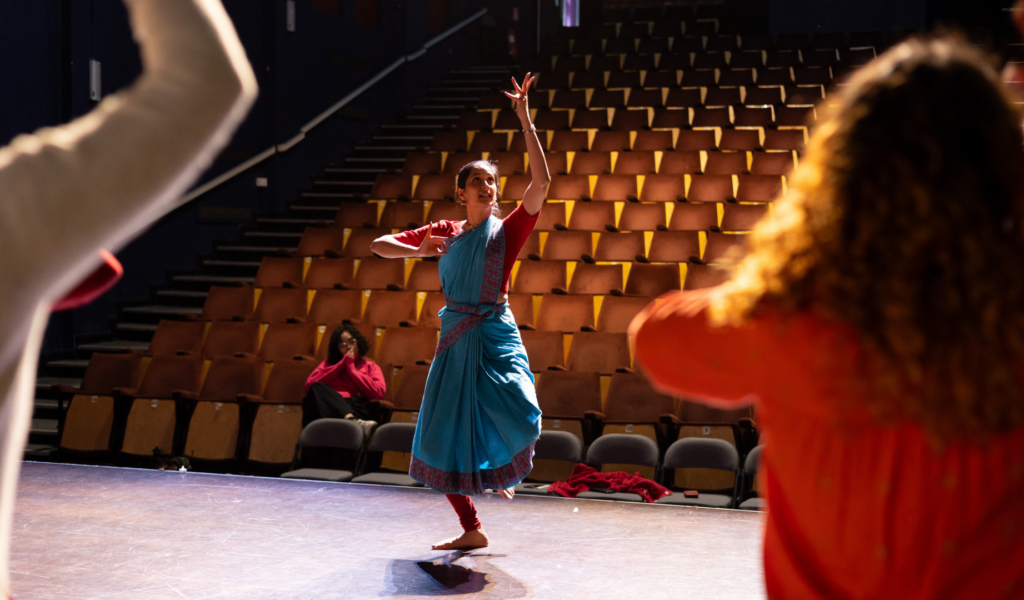 Photo of a woman in a colourful sari dancing on the Barnfield stage. Two people in the foreground copy her movements, with their backs to the camera.