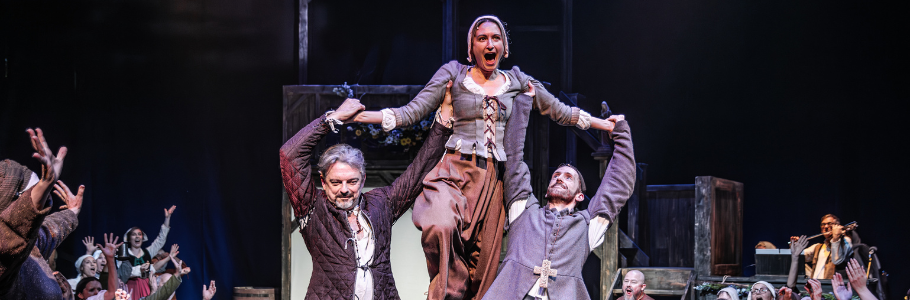 Photo of a woman being lifted over a fire by two men. They are surrounded by cheering ensemble members. All are in medieval dress and look joyful.