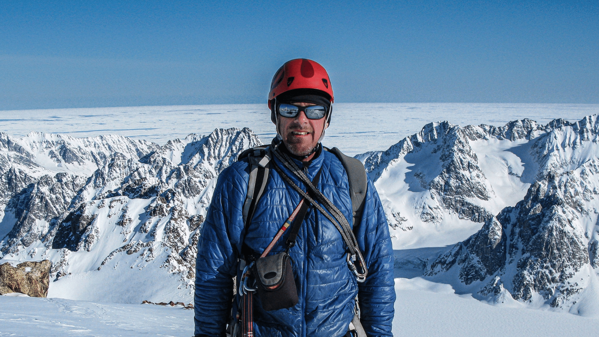 Simon Yates wearing a helmet and a winter jacket, standing in front of a snowy mountainous landscape .