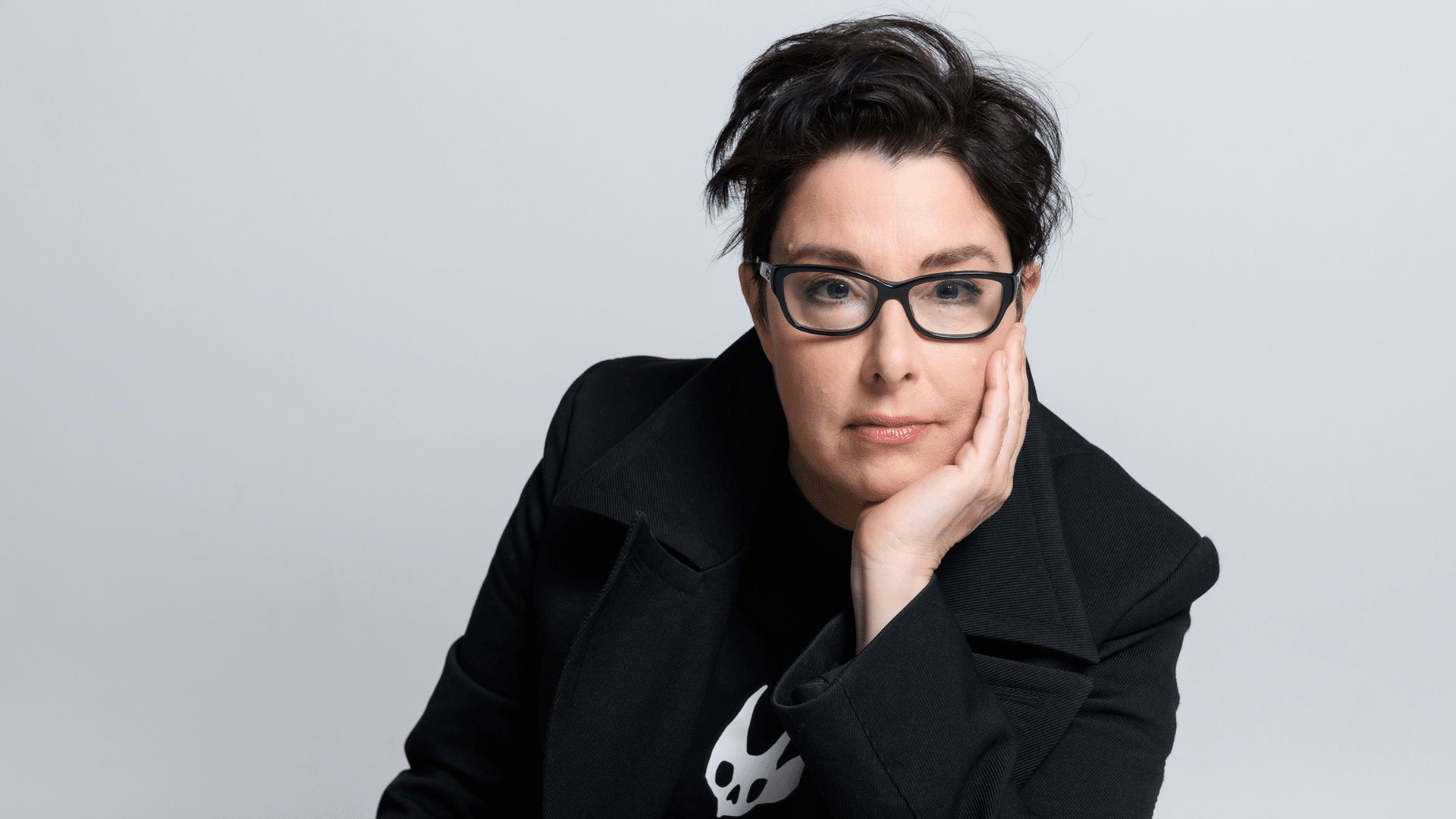 Sue Perkins posed in front of a white backdrop, resting their chin in the palm of their hand.