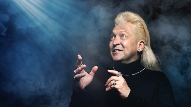 Photo of Clinton Baptiste wearing a black polo-neck and a white gold necklace and rings, with his long bottle-blond hair pushed back looking towards beams of light. The background is blue and smoky.