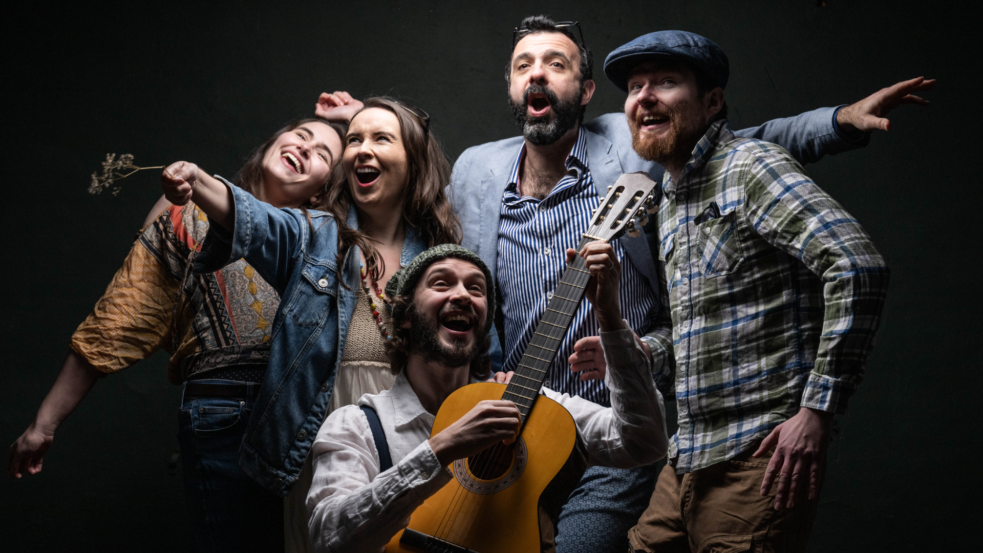 A photograph of a group of people in modern dress merrily singing and dancing together. At the front of the group crouches a man playing the guitar.