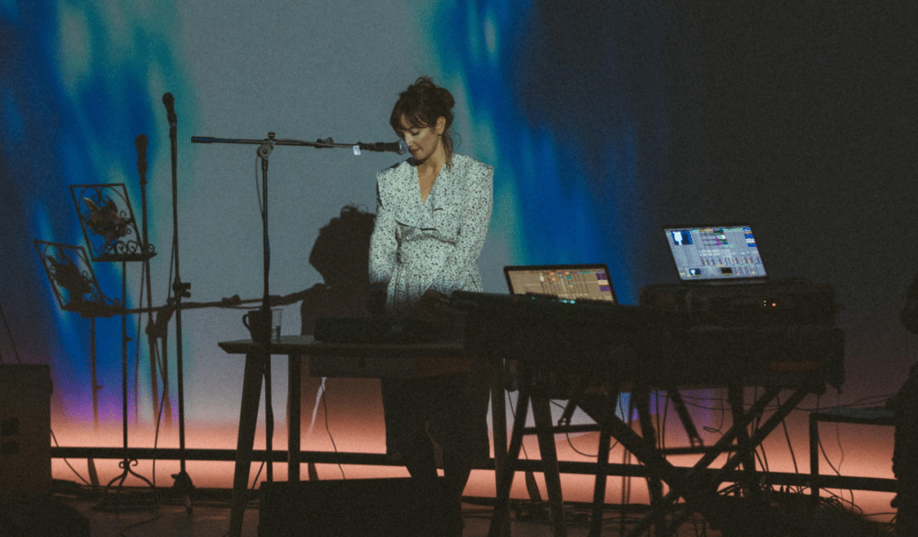 Photograph: a woman in a long floral dress stood behind two tables covered in sound equipment. They have a microphone positioned in front of them on a stand.