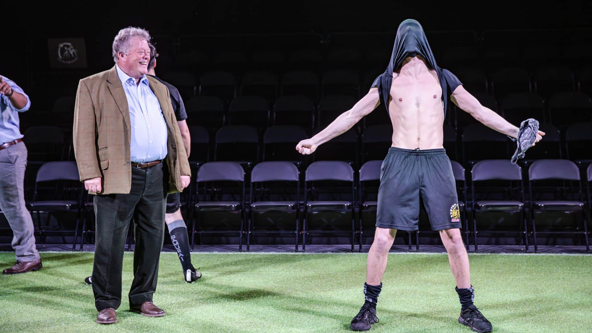 The character of Tony Rowe is looking at a young chiefs player who has pulled his shirt over his head and is celebrating bare-chested, laughing.
