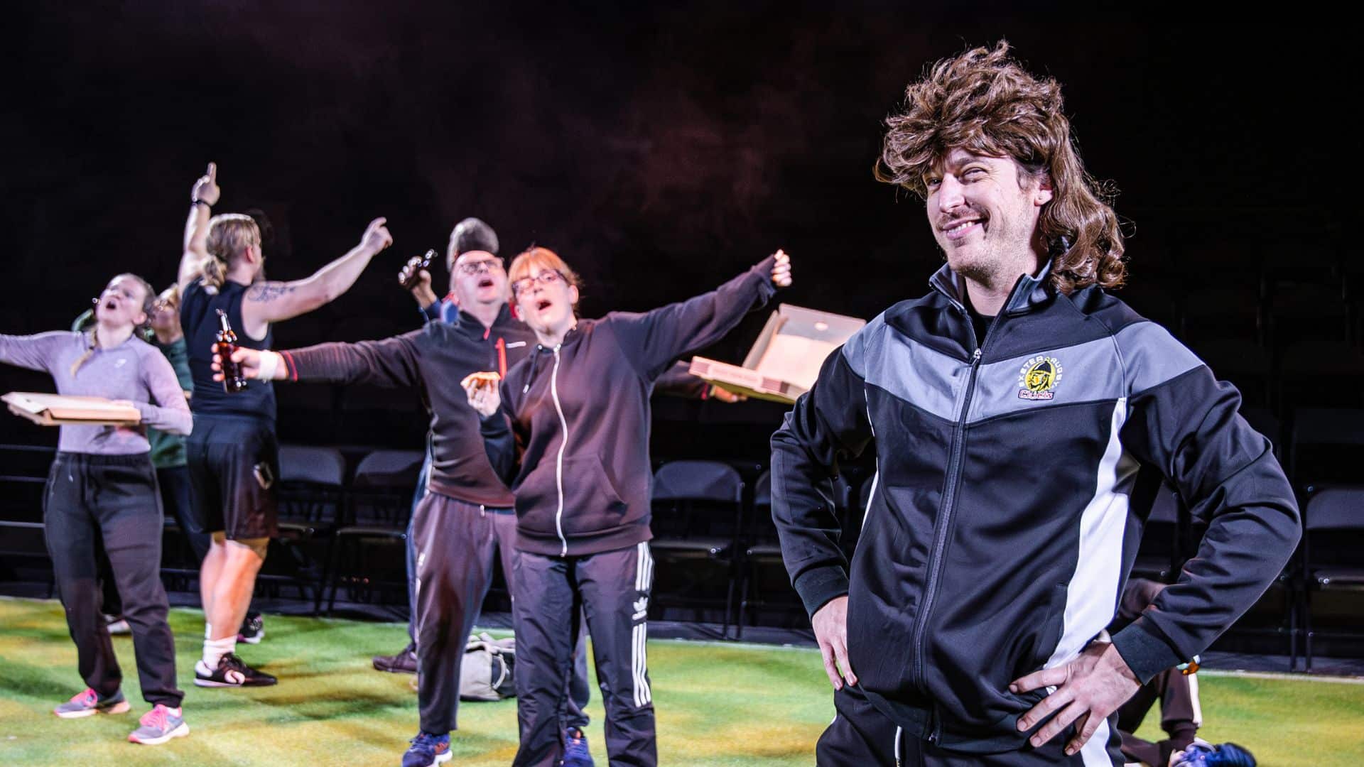 A Chiefs player with a funny mullet wig is grinning at the camera. In the background, others are having a party with beer and pizza.