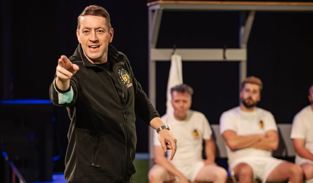 Exe Men production image. Robert Shaw Cameron playing Rob Baxter gives a motivational speech, pointing towards the audience. The cast as rugby players listen intently.