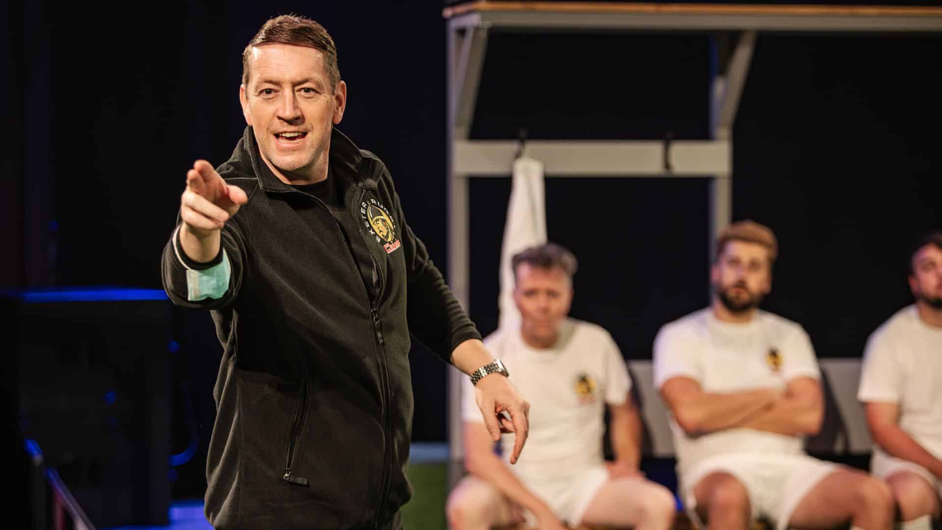 Exe Men production image. Robert Shaw Cameron playing Rob Baxter gives a motivational speech, pointing towards the audience. The cast as rugby players listen intently.