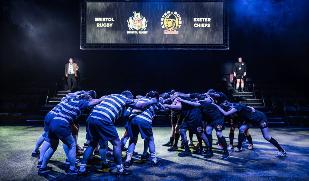 Exe Men production image. The Northcott stage has been transformed into Sandy Park rugby stadium. Players are in a scrum.