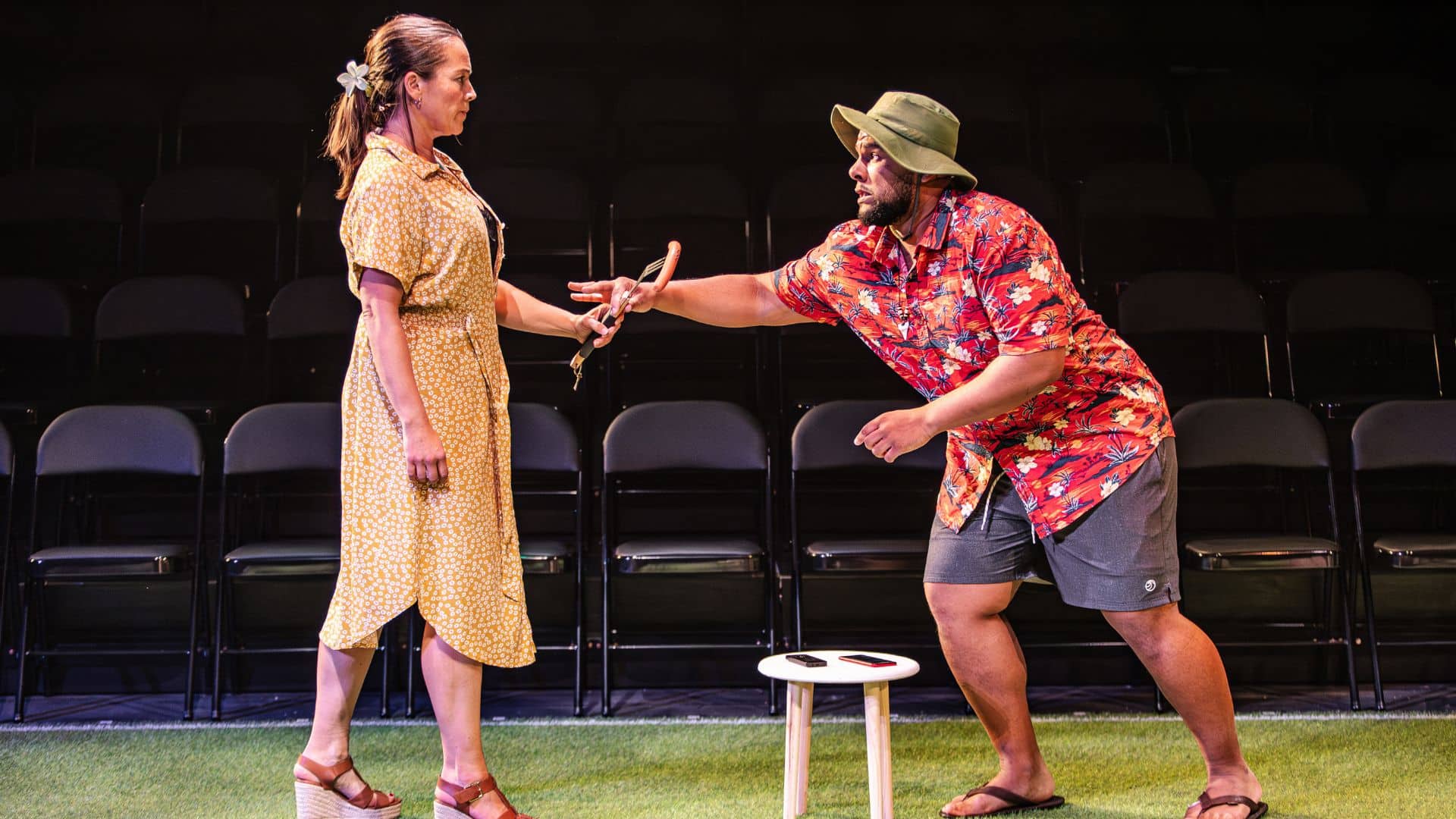 Exe Men production image. Micha Colombo and Emile Ruddock play a couple having a barbecue. Micha holds a sausage on a skewer, with a stern look on her face. Emile scrambles towards her with a worried look on his face.