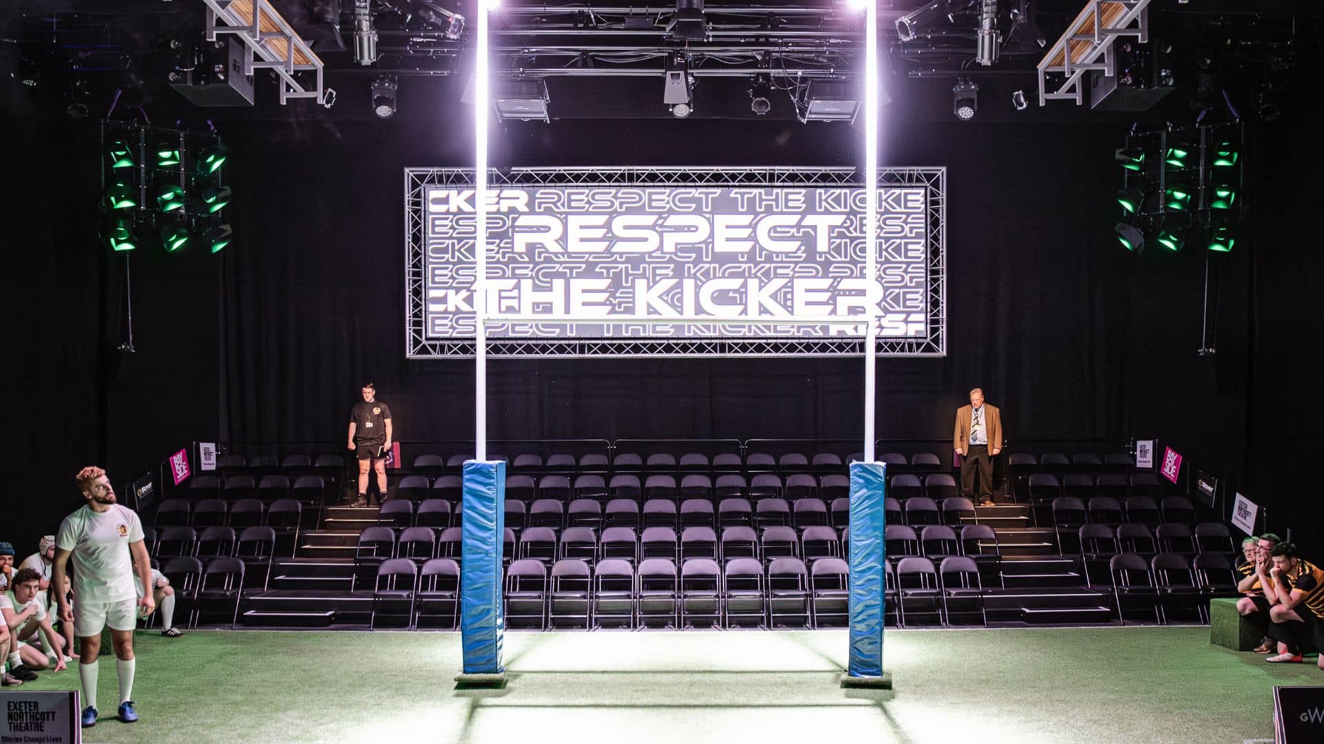 Exe Men production image. The Northcott stage has been transformed into Sandy Park rugby stadium. The cast gather on either side of the stage. Peter Watts stands looking at a big screen which reads 'RESPECT THE KICKER'.