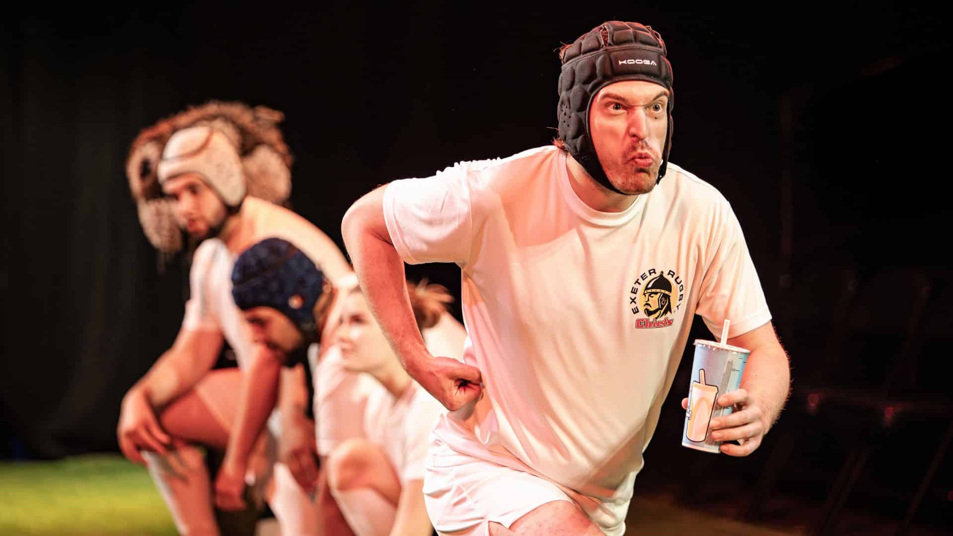 Exe Men production image. Joe Feeney runs as a rugby player. Other rugby players are crouched in the background.