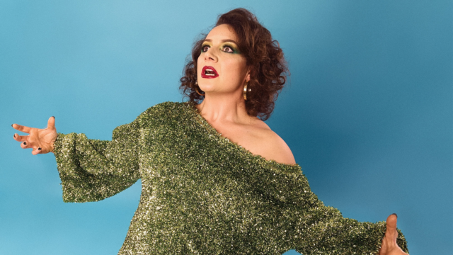 An image of Jessica Fostekew standing in shock looking upwards with her arms wide open. The background is a sky blue gradient and she's wearing a shimmery green top.