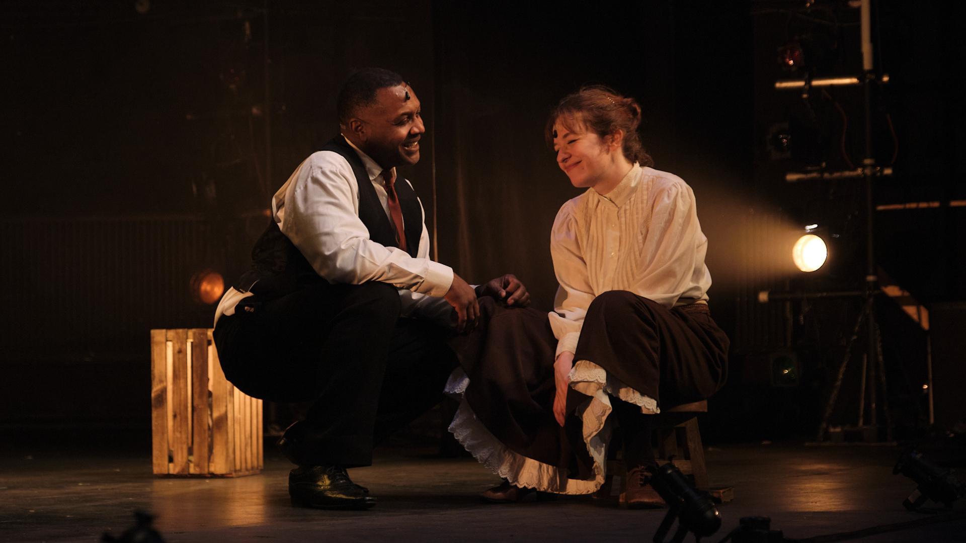 A production photograph of a man and woman crouching on stage smiling at each other.