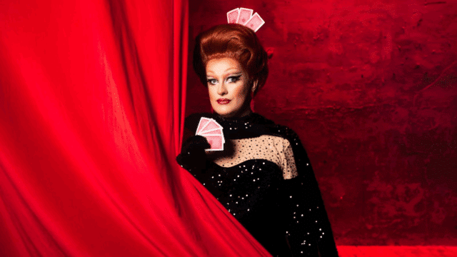 A photograph of Kate Butch appearing from behind a red curtain. She holds three playing cards in her right hand and has another three adorning in her wig.