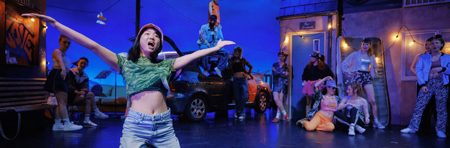 Photo of an actor on their knees with their arms spread wide and mouth open. In the background, a further twelve colourfully dressed actors look on, scattered around an urban street.