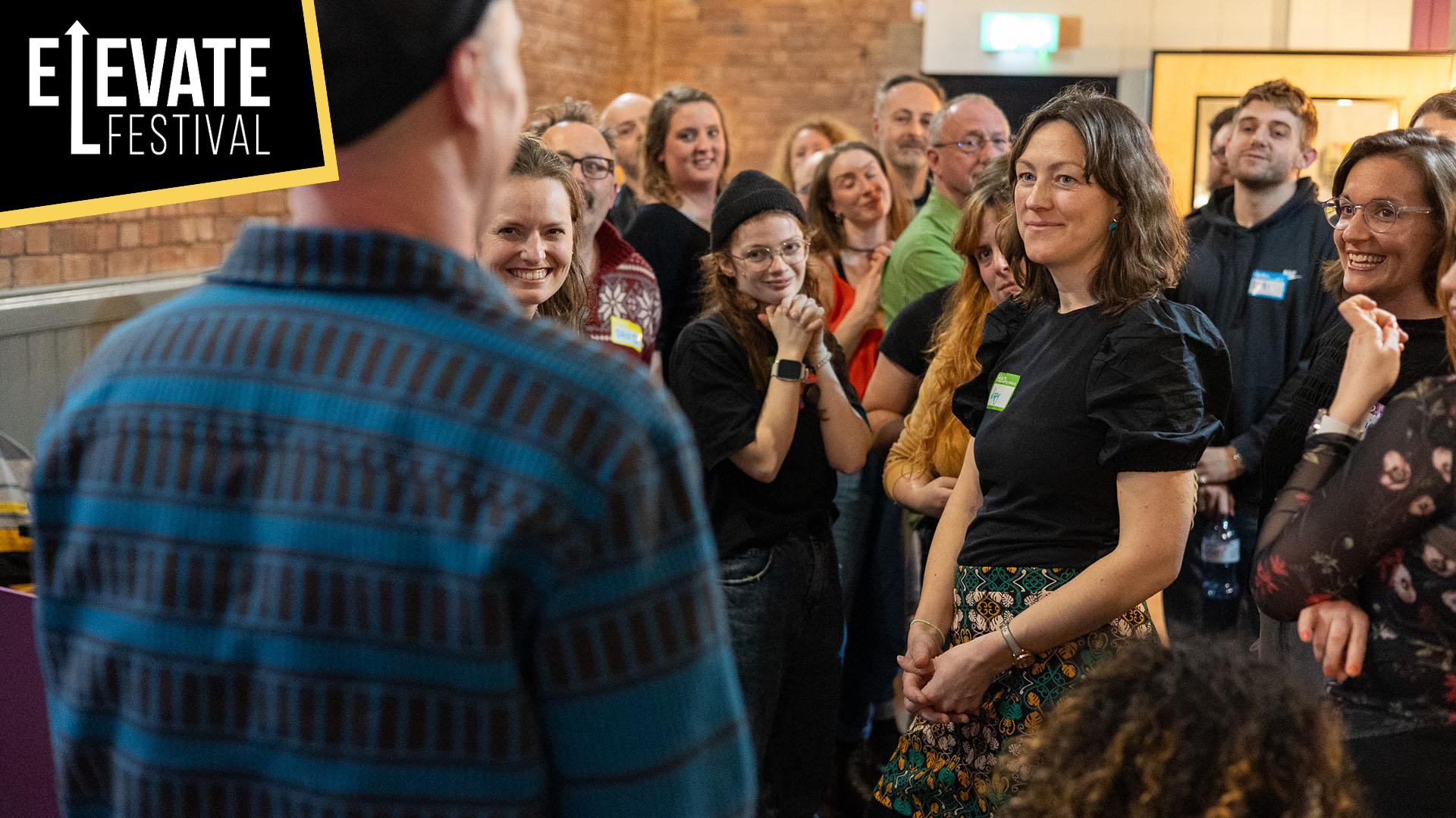 A group of Failspace participants stand and listen intently to another participant in the Barnfield Theatre's bar. text in the corner reads 'Elevate Festival'