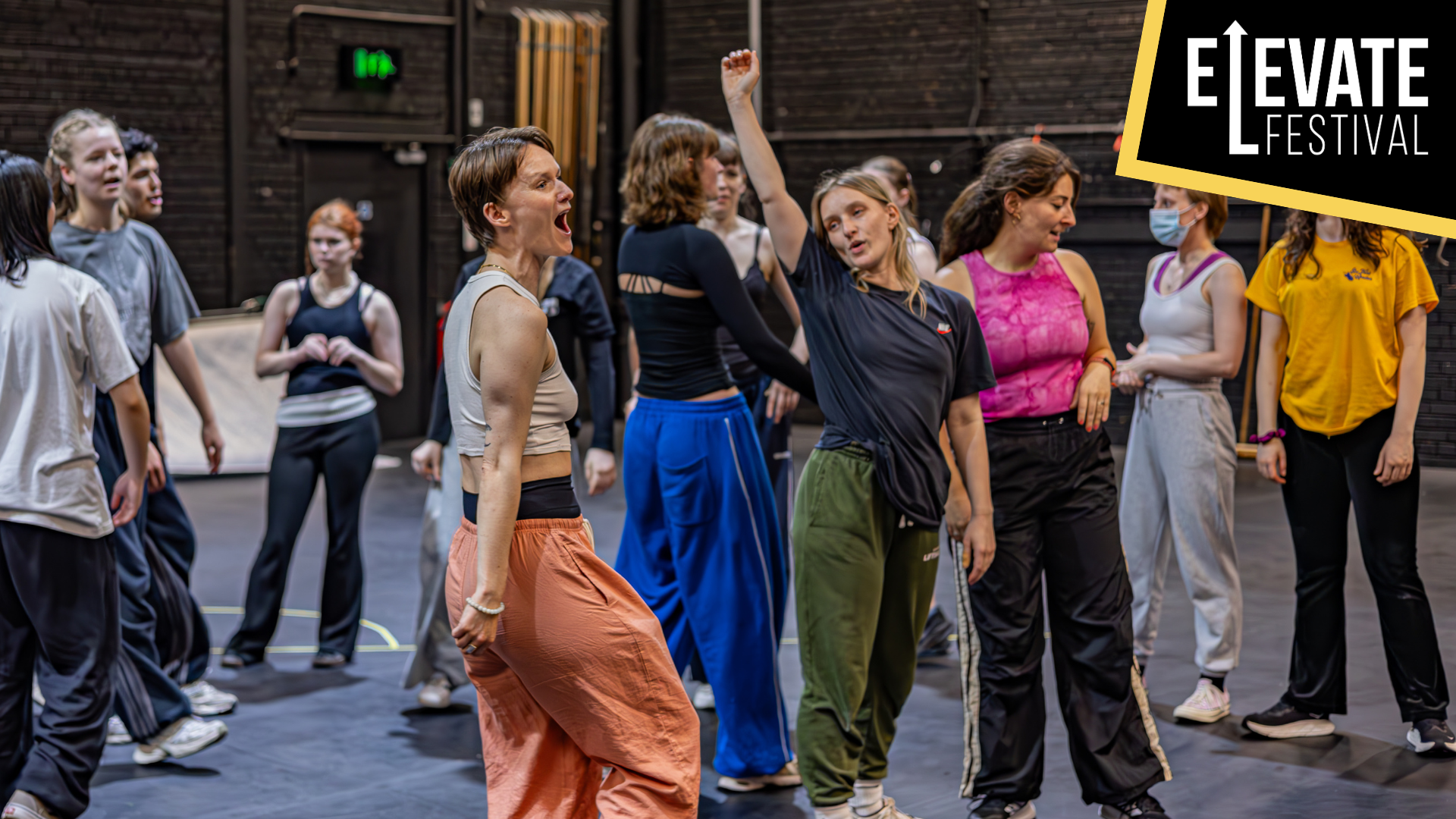 Photo of Romeo & Juliet rehearsals. A large cast, with two central figures: one screaming in excitement, the other raising one arm in a cheer. Elevate Festival logo.