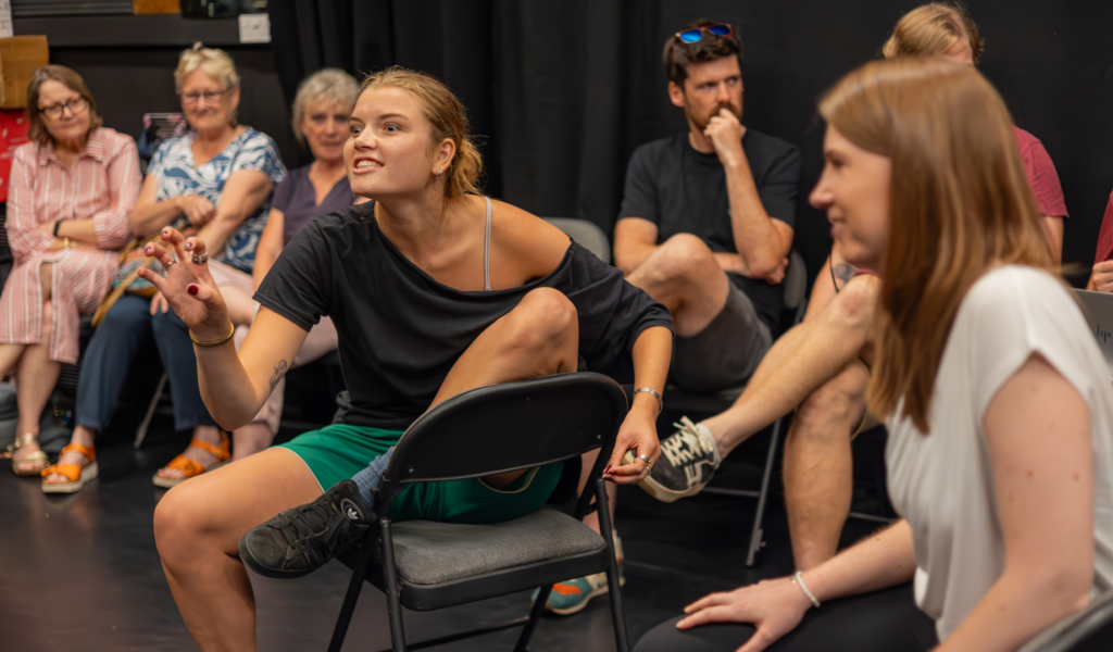 Exe Men rehearsal photo. A woman sits with one leg hooked up on a chair, forming her fist into a claw.