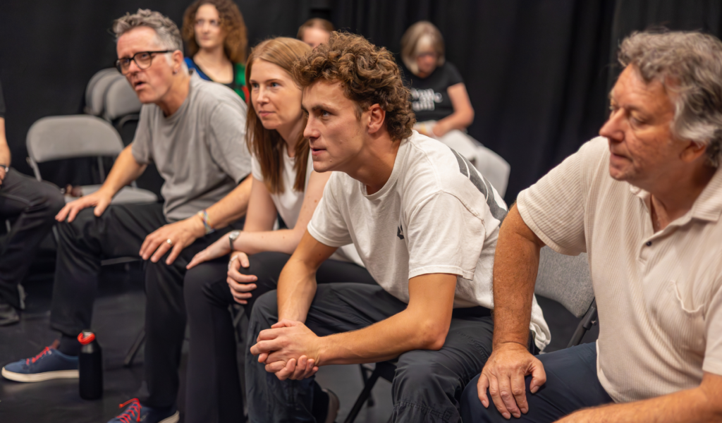 Exe Men rehearsal photo. Four actors sit side-by-side on chairs, listening intently and leaning forwards.