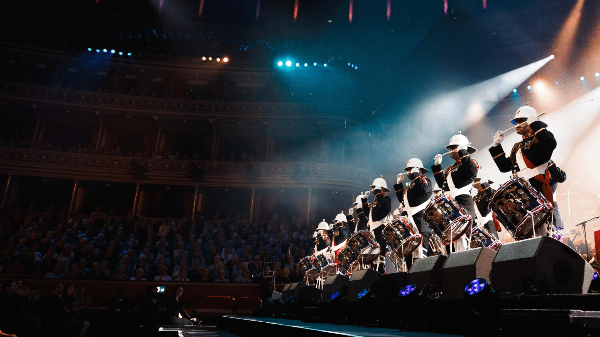 Photograph of a row of men on stage wearing military uniform and playing drums.