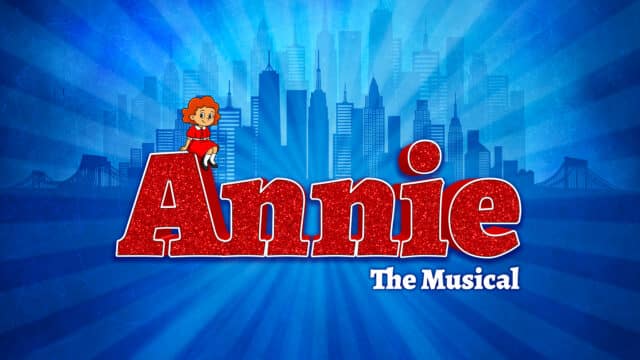 Annie The Musical promotional artwork: blue background with New York skyline. 'Annie' in large sparkly red letter with cartoon illustration of Annie sitting on the A.