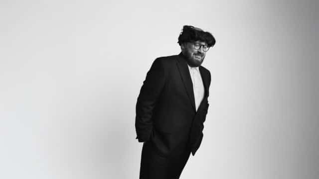 Black-and-white photo of John Kearns wearing a black suit and white shirt. He's leaning forward with his entire body straight. He appears to be wearing a silly wig that is bald in the centre and fake teeth.