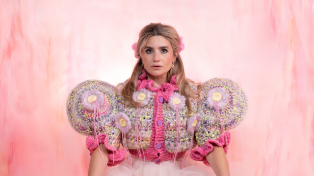A photograph of Harriet Kelmsley. She is stood in front of a light pink background wearing an elaborate pink knitted cardigan adorned with knitted rosettes.