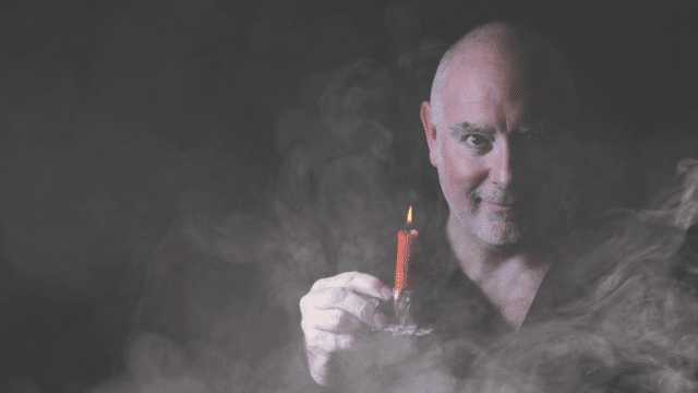 A photograph of an older man wearing a black shirt in a dark room. He holds a red candle as mist rises around him.