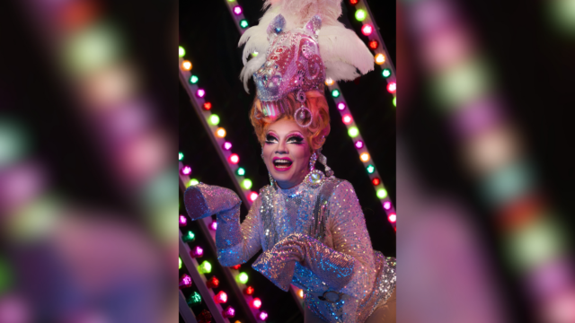 A photograph of Ginger Johnson in an extravagant sparkly carousel horse costume. Behind her are multicoloured fairground lights.