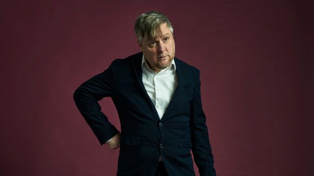 A photo of Tim Key staring wearily into the distance with a hand rested on his hip.