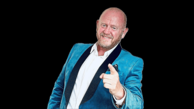 A photograph of John Smith against a black background. He is wearing a blue dinner jacket with black lapels and pointing at the viewer.