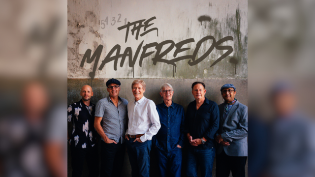 A photo of The Manfreds stood in a line in front of a derelict looking wall. Above them, written in spray paint, reads 'The Manfreds'.