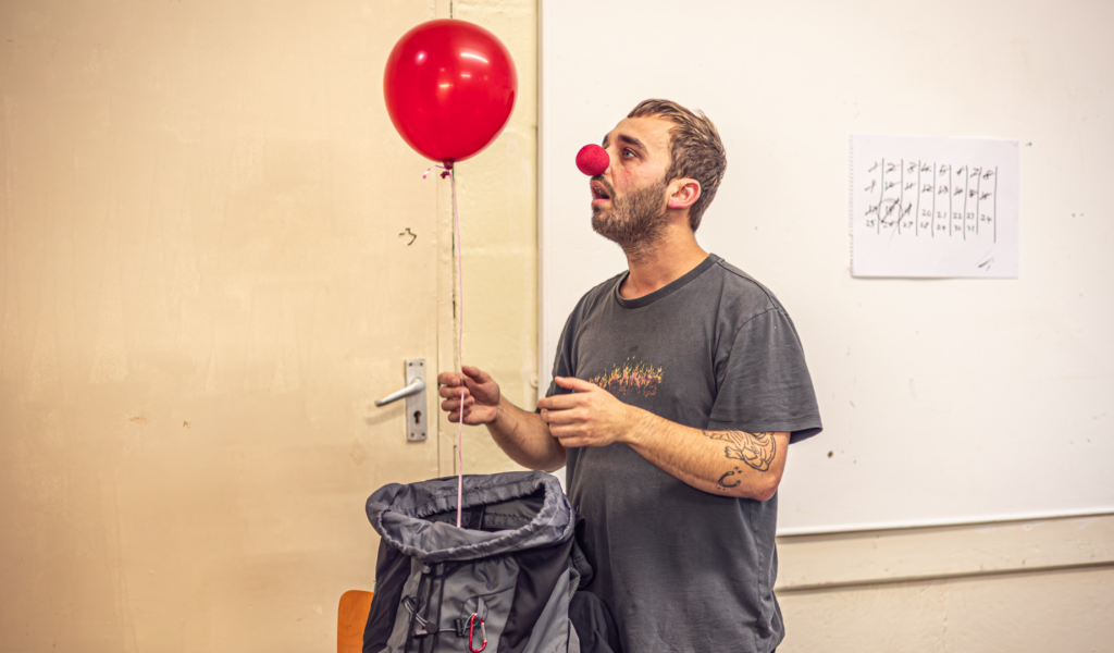 A man with a red nose looking at a red balloon coming out of a sack in front of him