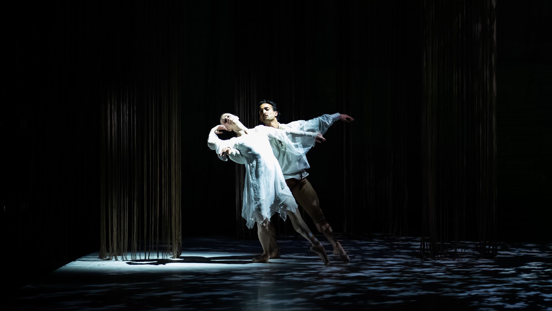 Two dancers wearing all white. The female dancer is in front with the right hand caressing her face. The male dancer is behind her his right arm under her arm reaching towards the audience and his left arm reaching out behind him.