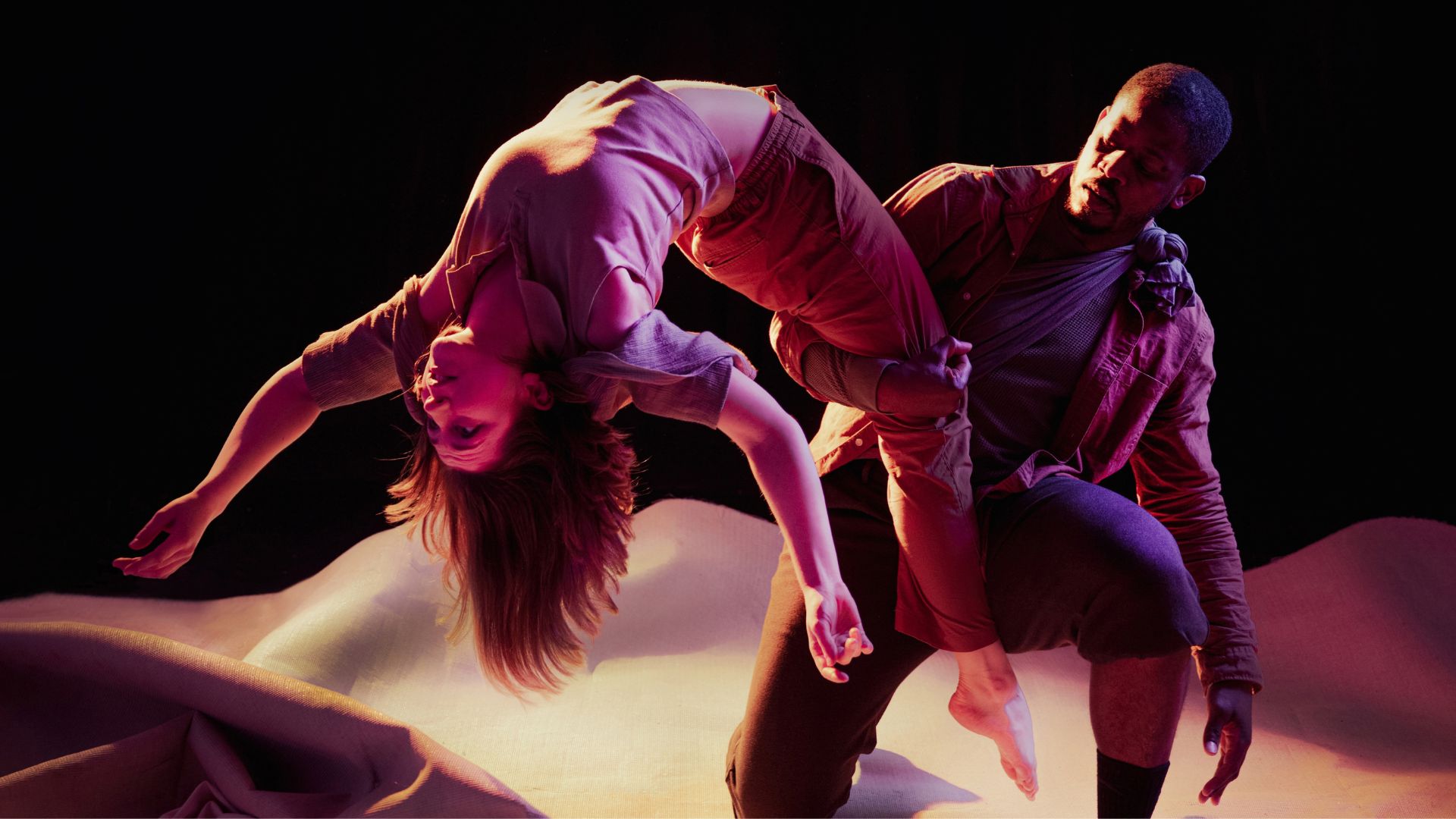 Two dancers hold a dramatic pose on stage, with the male dancer kneeling on one knee and supporting the female dancer by one leg as she arches backwards.
