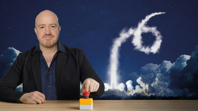 A photograph of Andy Parsons sitting at a desk, pressing a big red button. Behind him a rocket shoots into the the sky leaving a trail of smoke.