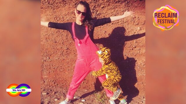 An image of a woman wearing a pink dungaree over a black t-shirt, standing with her arms wide open on brown dry gravel. A child wearing a tiger animal print onesie is holding onto her leg with their hands and legs. The top right corner has the Reclaim Festival logo and the bottom left corner has the We Are ND logo.