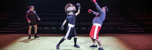 Exe Men production photo. Otter, the Chiefs rugby team's mascot, is wearing boxing gloves and is knocking out a caricaturistically French opponent.