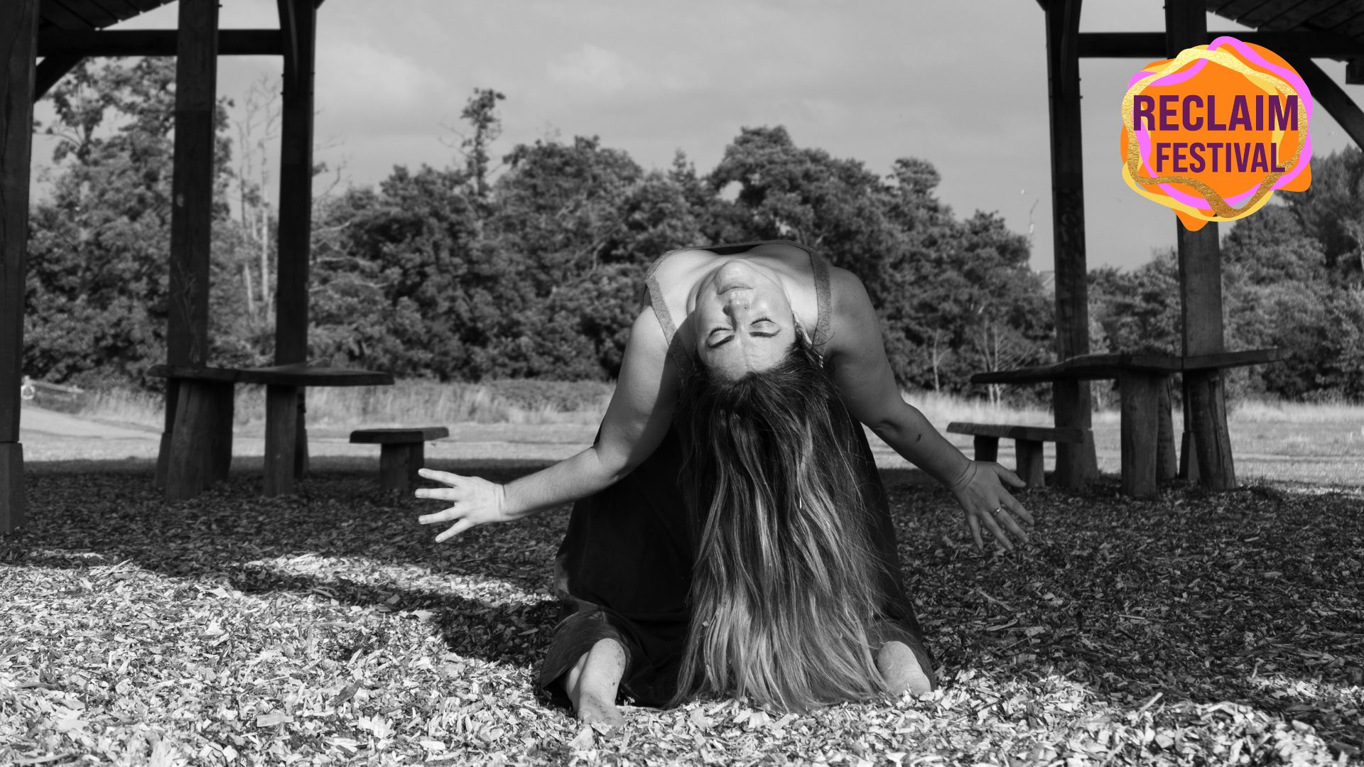 A black and white photograph of a woman dancing in a shady, covered area in a park. She is on her knees with her body arched back and her long hair hitting the floor behind her. The top right corner of the image has the RECLAIM FESTIVAL logo.