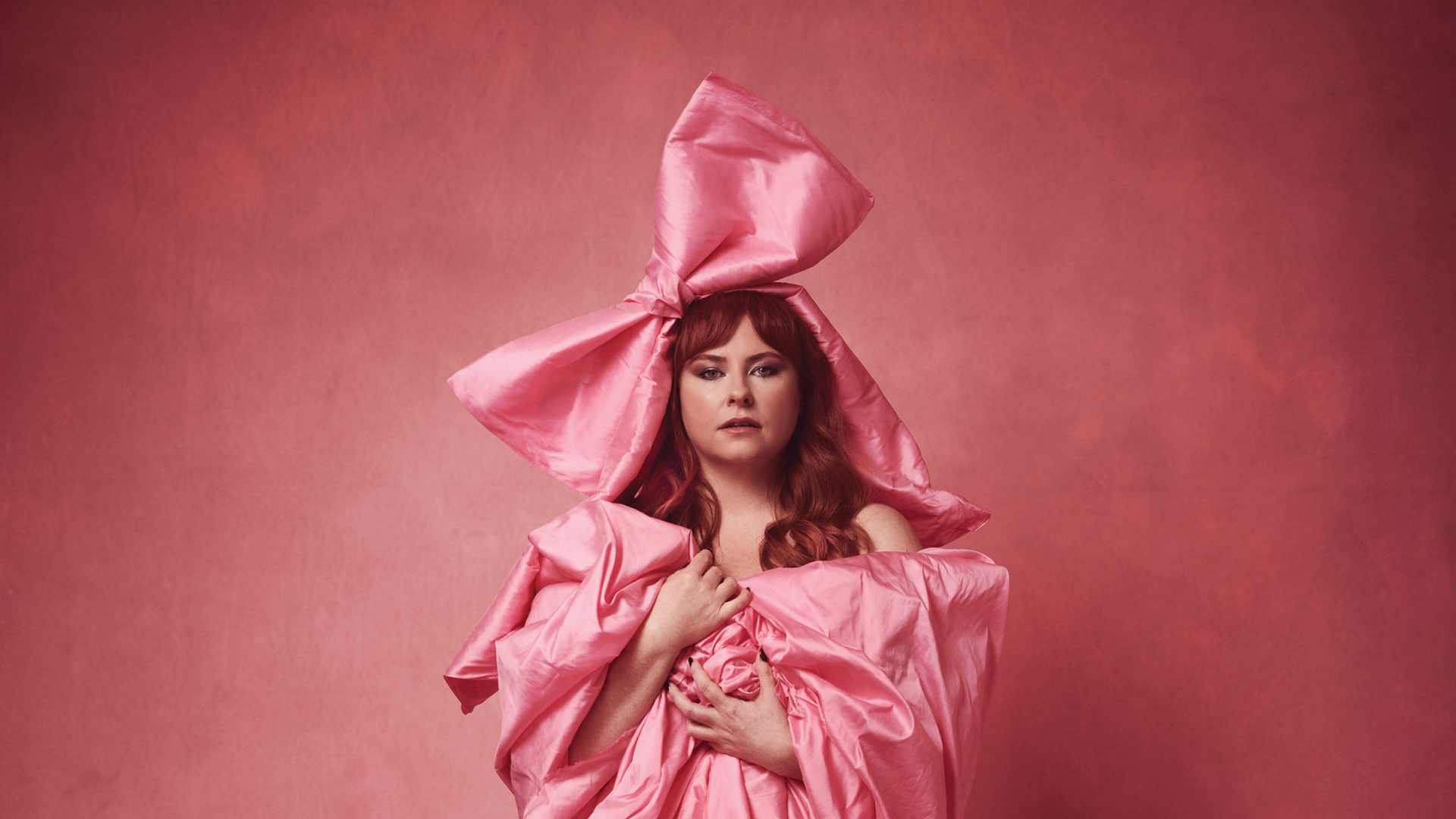 A photograph of Amy Gledhill against a pink background, wrapped in pink billowing satin fabric. She has an oversized bow on her head.