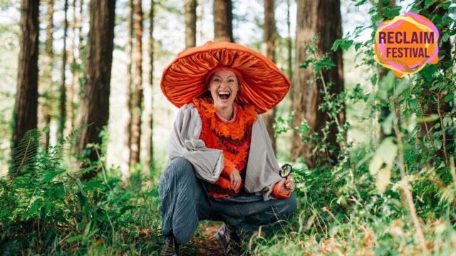 A person wearing a bright orange big mushroom hat is sitting down in the middle of trees in a forest. The top right of the image has the RECLAIM FESTIVAL logo.