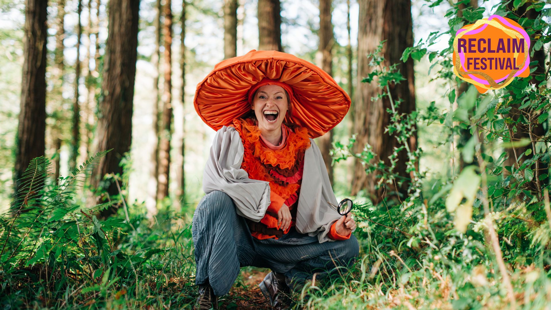 A person wearing a bright orange big mushroom hat is sitting down in the middle of trees in a forest. The top right of the image has the RECLAIM FESTIVAL logo.
