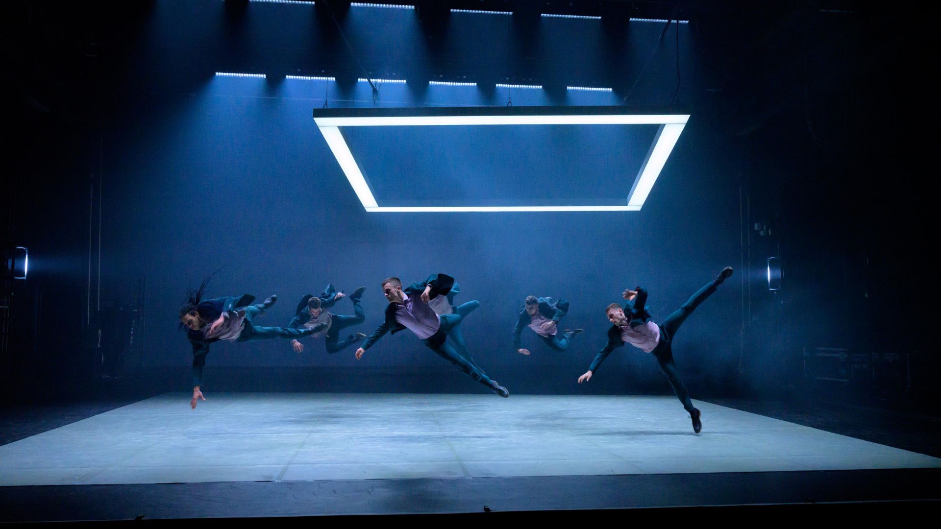 An image of Ballet Boyz performers on stage. They are all wearing suits and are doing a turn in the air in sync.