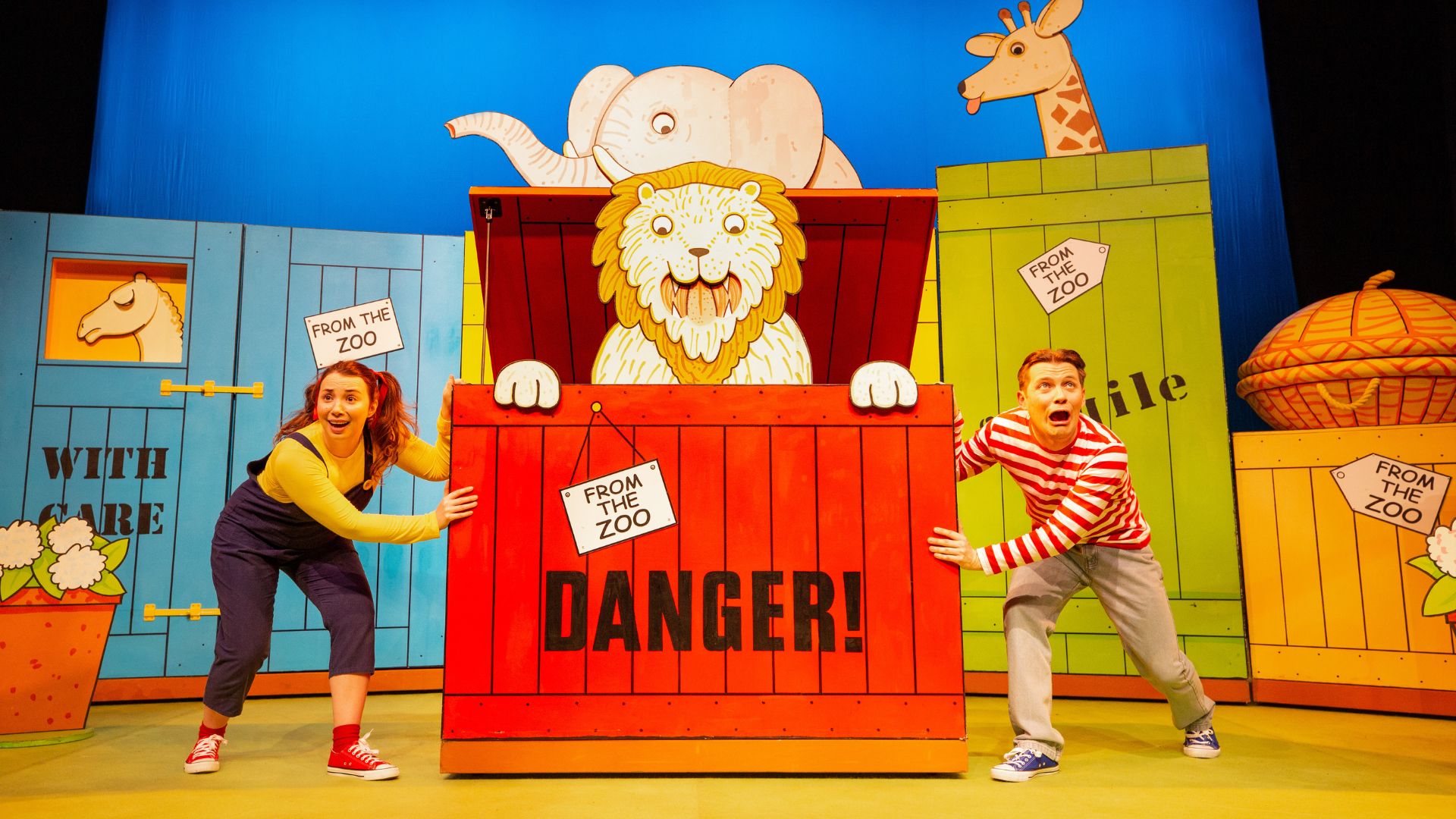 Dear Zoo live. Two actors on stage stood next to a bright red box with words DANGER written on it. A cardboard lion is spring out of the box.