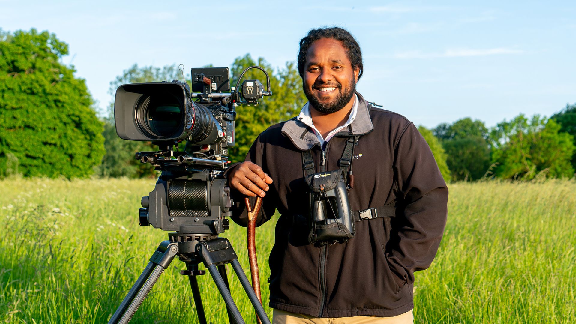 A photo of Hamza Yassin looking at the camera and smiling. He is standing in a field with his arm resting on a filming camera.
