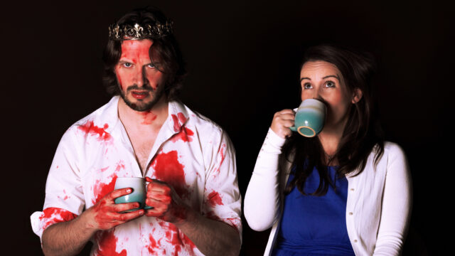 A man and a woman in front of a black screen. the man is wearing a crown, a white shirt covered in blood and holding a mug with an angry look on his face. Next to him is the woman wearing a blue dress and white cardigan sipping from her mug.
