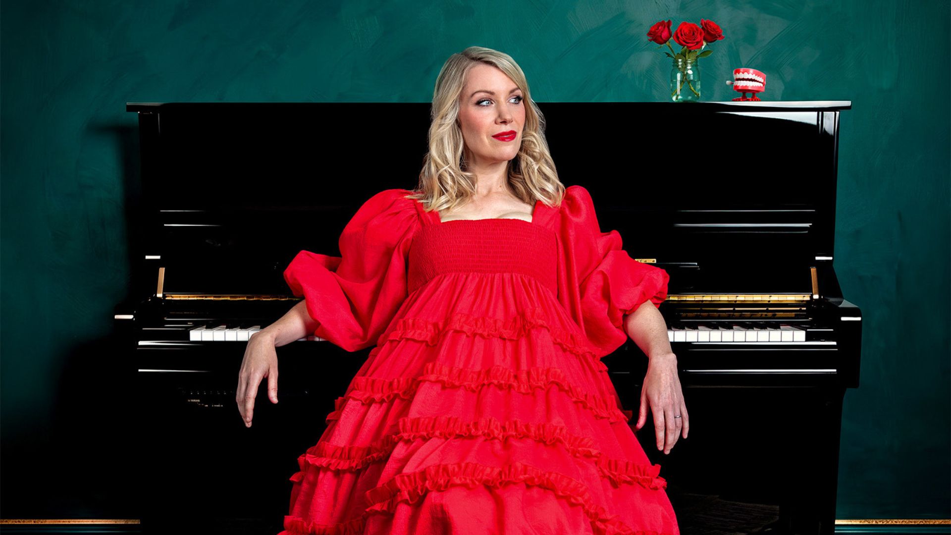 A photo of Rachel Parris looking away from the camera wearing a red long-sleeved dress with ruffles on the bottom. She is sitting in front of a piano with her elbows rested on the keys.